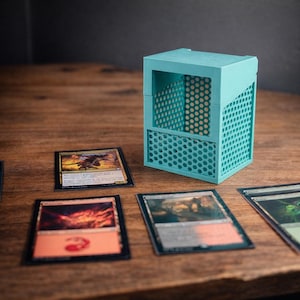 May include: A teal card storage box with a perforated design, next to several trading cards on a wooden surface. The box has a rectangular shape with an angled top and a visible interior.