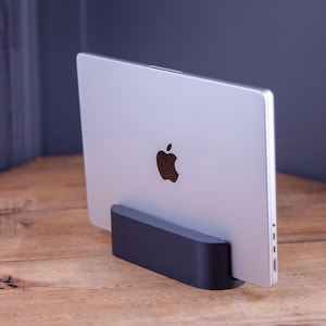 May include: A silver laptop with the Apple logo is displayed in a black stand on a wooden surface. The laptop is angled slightly, showcasing its sleek design. The stand provides support and elevates the device.