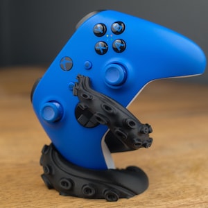 May include: A blue Xbox controller is held by a black octopus-tentacle-shaped stand. The controller has black buttons and a black directional pad. The stand is on a wooden surface.