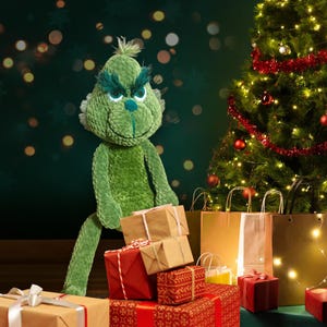 Puede incluir: Un juguete de peluche verde Grinch con una expresión gruñona está de pie junto a un árbol de Navidad y una pila de regalos envueltos. El Grinch sostiene una caja de regalo marrón en su mano.