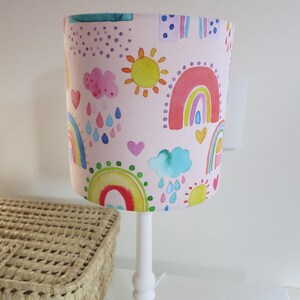 Könnte beinhalten: Eine weiße Lampe mit einem zylindrischen Lampenschirm mit einem Aquarelldesign aus Regenbogen, Sonnen, Wolken und Regentropfen auf rosa Hintergrund. Die Lampe steht auf einer weißen Oberfläche neben einem geflochtenen Korb.