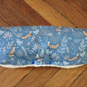 Könnte beinhalten: Ein blaues Kissen mit Fuchs-Motiv und einem weißen, flauschigen Rand. Das Kissen zeigt ein sich wiederholendes Muster aus Füchsen, Pflanzen und Blumen in Orange-, Weiß- und Rosatönen. Das Kissen ist rechteckig und hat blaue sternförmige Knöpfe.