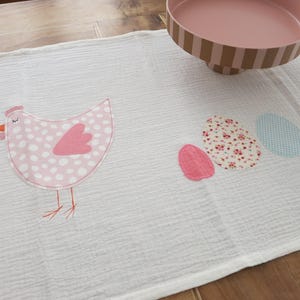 Könnte beinhalten: Weißer Tischläufer zum Thema Ostern mit einer rosa Henne und Eier-Applikationen. Die Henne hat weiße Tupfen und einen rosa Flügel. Die Eier sind rosa, floral und hellblau. Eine rosa-braune Servierschale befindet sich im Hintergrund.