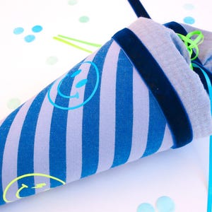 Könnte beinhalten: Ein Stoffkegel mit blau-weißen Streifen, grauem Oberteil und marineblauem Samtbesatz. Der Kegel hat ein Smiley-Gesicht-Design und ist mit blauen und grünen Bändern gebunden. Der Hintergrund ist weiß mit blauen und grünen Punkten.