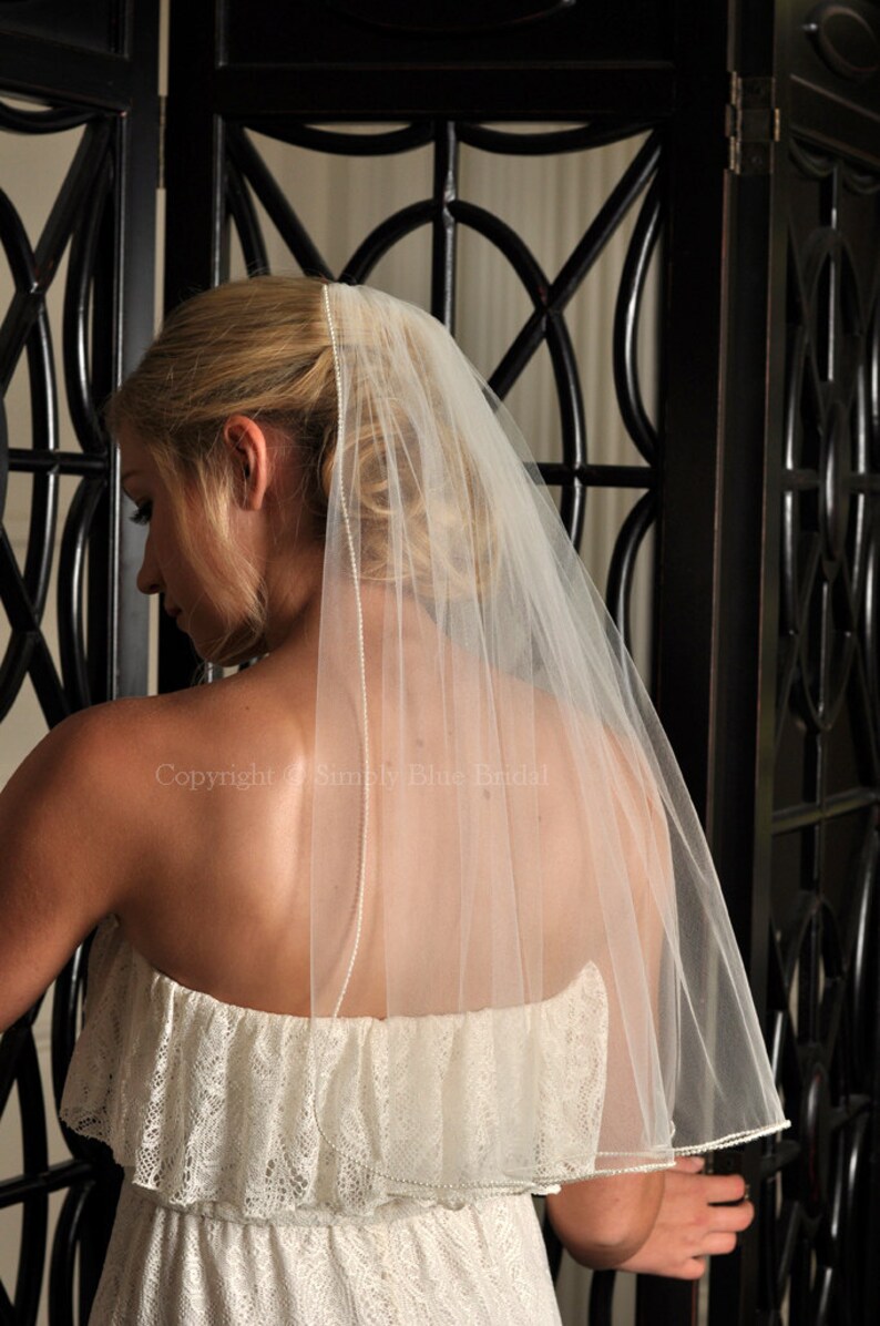 May include: A white bridal veil with a single row of pearls along the edge. The veil is made of soft tulle and is worn over a white lace dress.