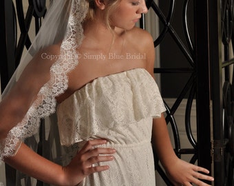 May include: A white lace wedding dress with a strapless bodice and a flowing skirt. The dress is paired with a white lace veil.