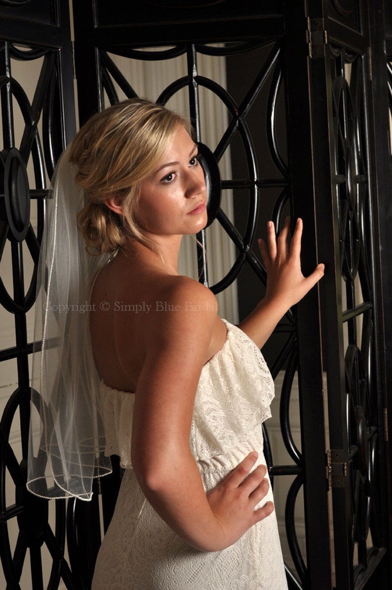 May include: A woman wearing a white lace wedding dress and a white veil with a beaded edge. She is standing in front of a black metal screen.