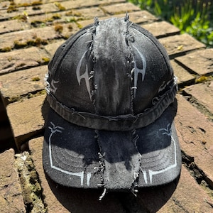 Op de afbeelding: Een zwarte baseballcap met witte ontwerpen in tribale stijl. De cap heeft een distressed look met zichtbare stiksels en gerafelde randen. De klep heeft ook een wit ontwerp. De cap ligt op een bakstenen oppervlak.