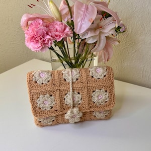 May include: A crocheted pouch with a floral design in shades of peach and cream. The pouch has a tassel and is made with a granny square pattern.