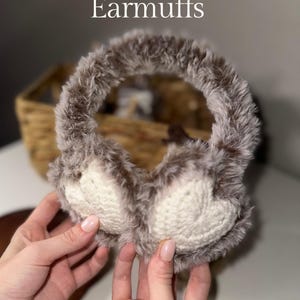 May include: Crochet pattern for fuzzy heart earmuffs. The earmuffs are made with brown faux fur and have white crocheted hearts on the front. The earmuffs are being held by a person's hands.