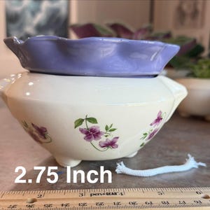 May include: A cream-colored ceramic planter with a ruffled lavender rim and purple floral decals. A white wick rests in front of the planter, which measures 2.75 inches in height as indicated by the visible ruler at the base.