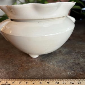 May include: A cream-colored ceramic planter with a ruffled rim and three small feet. The planter has a rounded body and a slightly flared top. The ruler next to the planter shows measurements in inches.