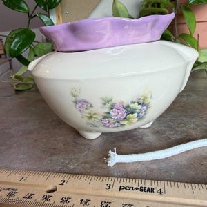 May include: A cream-colored ceramic bowl with a lavender rim and floral design. The bowl has three small feet and a decorative floral pattern in shades of purple and green. A white rope is next to the bowl.