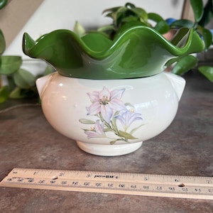 May include: A two-piece ceramic bowl with a green, ruffled top and a white base. The base features a floral design with pink and purple lilies. A ruler is placed near the bowl, measuring up to 12 inches.