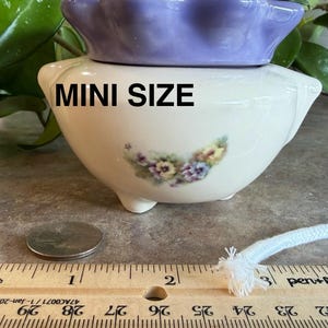 May include: A cream-colored ceramic planter with a lavender ruffled rim and a floral decal on the front. The text MINI SIZE is printed in black. A ruler shows the planter is approximately 3 inches wide, placed next to a coin and a white wick.