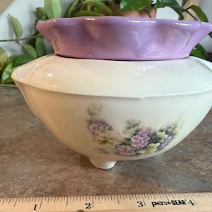 May include: A two-piece ceramic set featuring a cream-colored bowl with a floral design and a smaller, lavender-colored bowl on top. The larger bowl has three small feet. The floral design includes pink and green flowers.