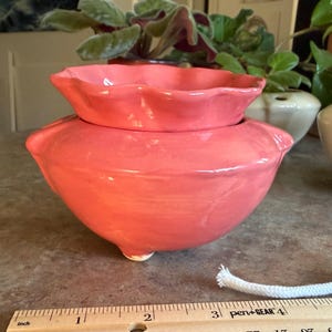 May include: A coral-colored ceramic planter with a ruffled top edge and a rounded base, featuring a small white wick extending from the bottom. A wooden ruler is positioned in the foreground, showing the planter is approximately 4.5 inches wide.