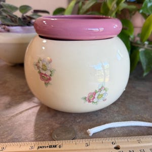 May include: A round, cream-colored ceramic planter with a smooth, glossy finish and a thick, solid pink rim. The body features small, delicate floral clusters in shades of pink, white, and green. A wooden ruler is visible at the base for scale.