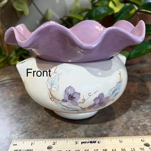 May include: A two-piece ceramic decorative bowl. The top piece is a wavy-edged lavender bowl. The bottom piece is a white bowl with a floral design in purple and blue. The word "Front" is visible on the bowl.