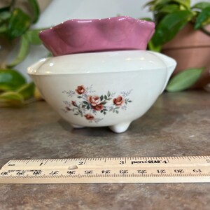 May include: A two-piece ceramic container with a white base and a pink, scalloped lid. The base features a floral design with red roses and green leaves. The container is approximately 4 inches wide.