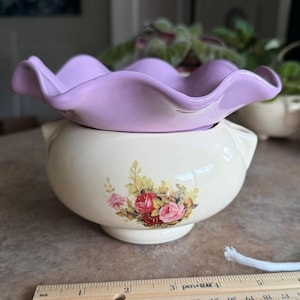 May include: A cream-colored ceramic bowl with a floral rose decal and a removable lavender ruffled top. A wooden ruler placed in front shows the bowl is approximately 6 inches wide. The bowl features small side handles and a pedestal base.