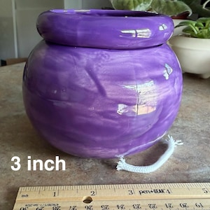 May include: A round, glossy purple ceramic self-watering planter with a smooth finish and a thick, rounded rim. A white cotton wick extends from the base. A wooden ruler placed in front shows the planter has a diameter of 3 inches.