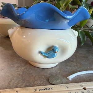 May include: A cream-colored ceramic bowl with a ruffled blue rim and a small blue bird figurine attached to the side. A metal coin and a white cord are placed near a wooden ruler that shows the bowl is approximately 5.5 inches in width.