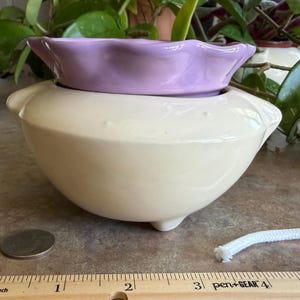 May include: A two-piece ceramic self-watering planter featuring a cream-colored base with three small feet and a removable lavender-colored top section with a scalloped rim. A white cotton wick is included for the self-watering mechanism.