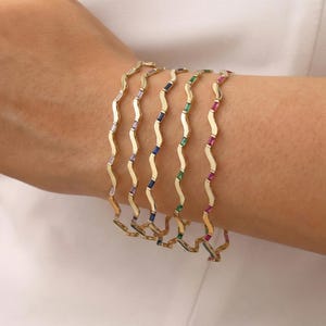 Può includere: Una collezione di braccialetti dorati ondulati. Ogni braccialetto è adornato con una fila di pietre preziose colorate, tra cui bianco, viola, blu, verde e rosso. I braccialetti sono mostrati su un braccio.
