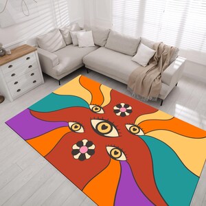 Può includere: Un tappeto rettangolare con un design retrò. Il tappeto presenta un motivo di occhi e fiori stilizzati nei toni dell'arancione, giallo, verde acqua, viola e marrone. Il tappeto è posizionato su un pavimento in legno chiaro in un soggiorno.