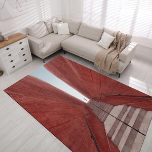 Puede incluir: Una alfombra abstracta roja y blanca con un diseño de una escalera que conduce a una abertura de luz. La alfombra está sobre un suelo de madera claro en una habitación con un sofá blanco y una cómoda blanca.