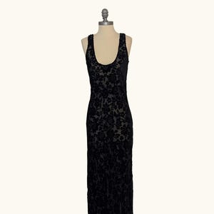 K&ouml;nnte beinhalten: Ein schwarzes, bodenlanges Kleid mit floralem Muster. Das Kleid hat einen Rundhalsausschnitt und ist &auml;rmellos. Der Stoff scheint Samt oder ein &auml;hnliches Material mit dezentem Glanz zu sein. Das Kleid wird auf einer Schaufensterpuppe pr&auml;sentiert.