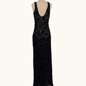 K&ouml;nnte beinhalten: Ein schwarzes, bodenlanges Kleid mit floralem Samtmuster. Das &auml;rmellose Kleid hat einen tiefen V-Ausschnitt am R&uuml;cken und wird auf einer Schaufenquin pr&auml;sentiert. Das Kleid ist aus einem weichen, flie&szlig;enden Stoff gefertigt.