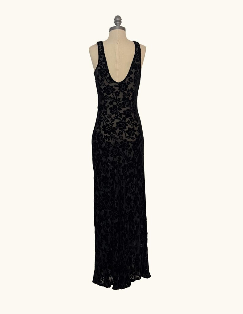 K&ouml;nnte beinhalten: Ein schwarzes, bodenlanges Kleid mit floralem Samtmuster. Das &auml;rmellose Kleid hat einen tiefen V-Ausschnitt am R&uuml;cken und wird auf einer Schaufenquin pr&auml;sentiert. Das Kleid ist aus einem weichen, flie&szlig;enden Stoff gefertigt.