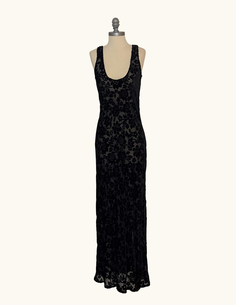 K&ouml;nnte beinhalten: Ein schwarzes, bodenlanges Kleid mit floralem Muster. Das Kleid hat einen Rundhalsausschnitt und ist &auml;rmellos. Der Stoff scheint Samt oder ein &auml;hnliches Material mit dezentem Glanz zu sein. Das Kleid wird auf einer Schaufensterpuppe pr&auml;sentiert.