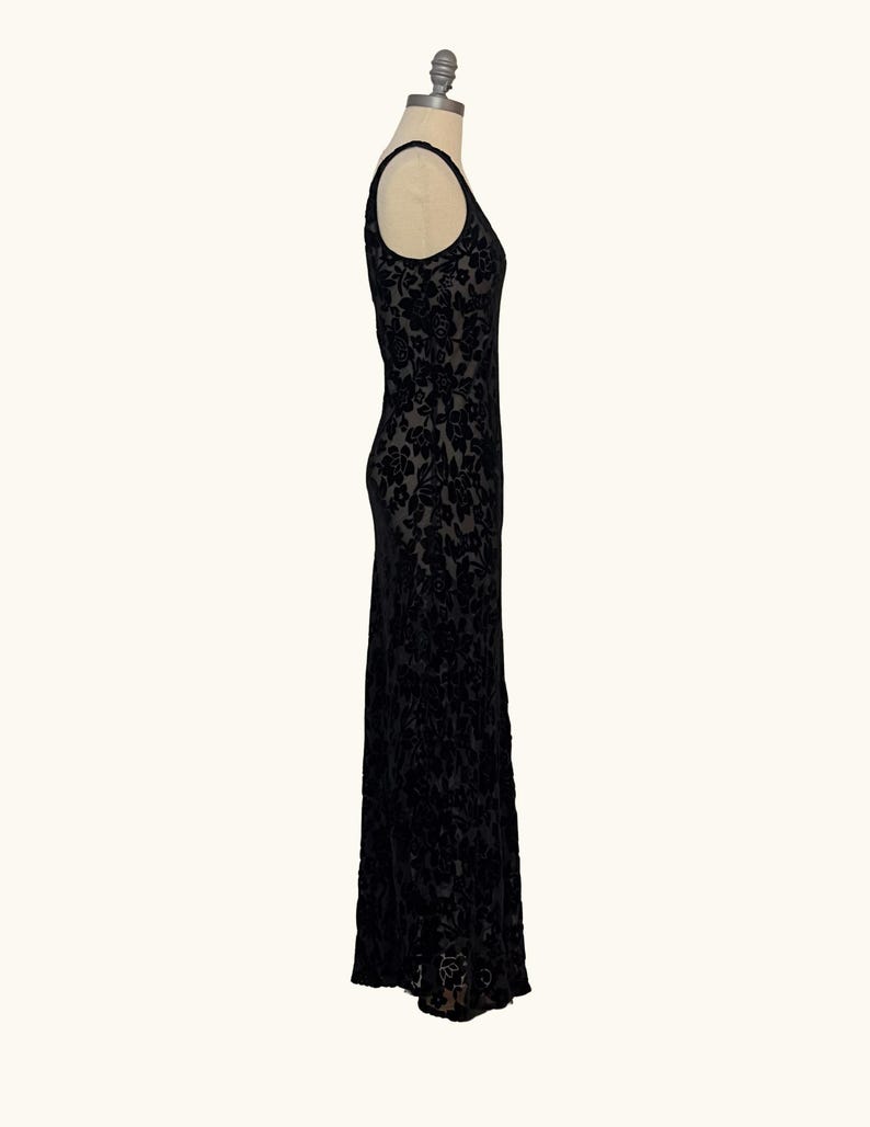 K&ouml;nnte beinhalten: Ein schwarzes, bodenlanges Kleid mit floralem Muster. Das &auml;rmellose Kleid wird auf einer Schaufensterpuppe pr&auml;sentiert. Der Stoff scheint Samt oder ein &auml;hnliches Material mit dezentem Glanz zu sein. Das Kleid hat einen Rundhalsausschnitt und eine taillierte Silhouette.