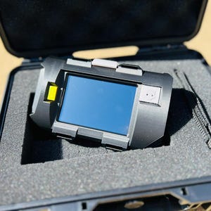 Op de afbeelding: Een zwart elektronisch apparaat met een blauw scherm, gele accenten en een koord, rustend in een open zwarte koffer met grijze schuimvulling. Het apparaat is het centrale punt van de afbeelding.