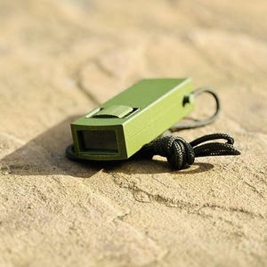 May include: A green rectangular whistle with a black lanyard rests on a textured, light brown surface. The whistle features a sliding mechanism and a small opening. The lanyard is knotted.