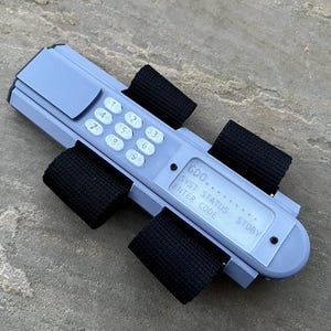 May include: A gray, rectangular device with a keypad and a display screen. The device has black straps attached to it. The display screen shows the text "GDO... SYST STATUS: STOBY ENTER CODE:"