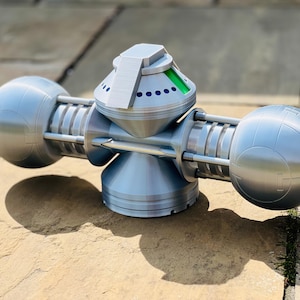 May include: A silver-colored, 3D printed model of a futuristic device with two spherical ends and a central, rotating section. The central section has a green and silver top with a small, silver lever.