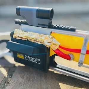 May include: Close-up of a futuristic device with a gray scope and black housing. The device has a yellow section with red tubing and is wrapped in gold foil. A caution label is visible.