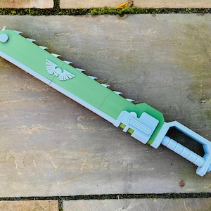 May include: A green and light blue toy chainsaw with a toothed blade and a handle. The chainsaw has a white emblem with a skull and wings. The toy is lying on a stone surface.