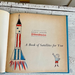 Puede incluir: Libro abierto con una cubierta azul y páginas envejecidas. La ilustración muestra un cohete con rayas rojas, blancas y azules, y un niño de dibujos animados señalando hacia arriba. El texto dice "A Book of Satellites for You."