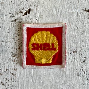 Puede incluir: Un parche cuadrado con el logotipo de Shell. El logotipo es una concha amarilla con la palabra "SHELL" en rojo. La concha está sobre un fondo rojo, con un borde blanco.