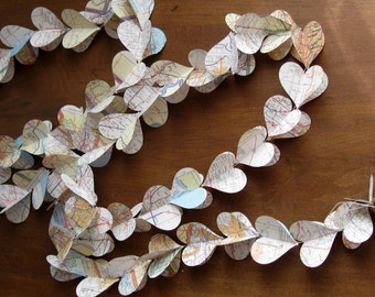 May include: A garland of paper hearts, each cut from vintage maps, lies on a wooden surface. The hearts are connected in a chain, creating a decorative element. The maps feature various colors and details, adding visual interest to the handmade craft.