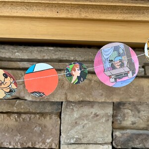 May include: A garland of circular cutouts with cartoon illustrations. The images include a bear, a character in a car, a profile of a person, a red and blue design, and a cartoon dog. The garland is strung on a white string.
