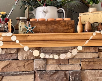May include: A decorative mantelpiece with a stone facade. The mantel displays a variety of items, including a garland of white circles, a painting, vases with flowers, jars, and a white teapot with a star design.