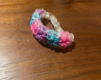 May include: A rainbow loom bracelet made with pink, blue and purple rubber bands. The bracelet is made with a clear rubber band base.