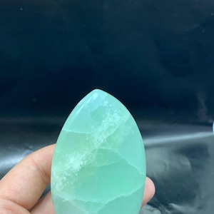 May include: A polished, oval-shaped gemstone in shades of light green. The stone has a smooth surface with visible internal patterns and a slightly translucent appearance. The gemstone is held in a hand, with a dark background.