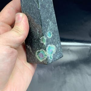 Può includere: Un obelisco di cristallo rettangolare grigio scuro con macchie di colore turchese e viola. Il cristallo è tenuto in mano, mostrando la sua forma geometrica lucida e i modelli minerali naturali. È un oggetto decorativo.